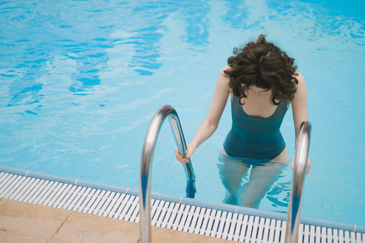 why-choose-us Young woman in a teal swimsuit using ladder to enter the pool on a sunny day.